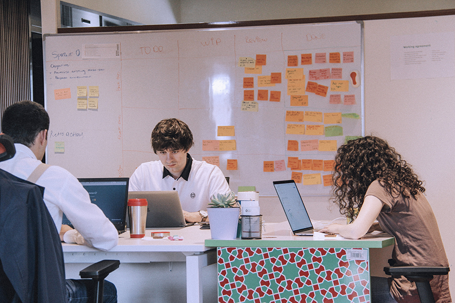 dois homens e uma mulher trabalhando em suas mesas com notebooks e atrás a imagem de uma lousa com vários post it colados